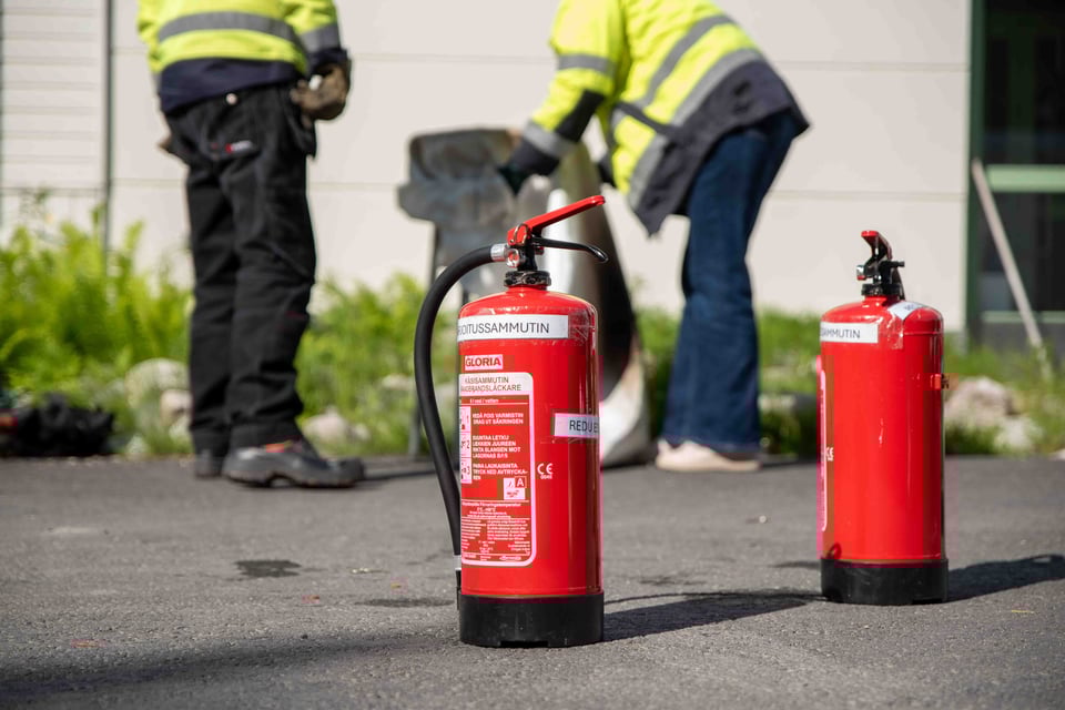 Punainen käsisammutin etualalla ja kaksi henkilöä epäterävänä taustalla suoja-asuissa tekemässä sammutusharjoitusta.