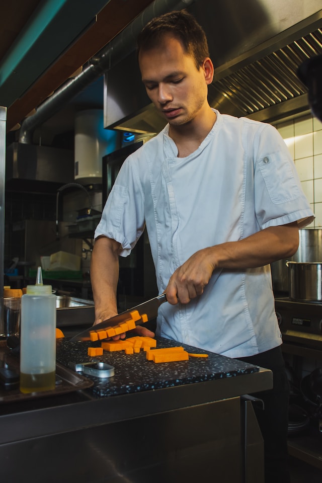 Keittiötyöntekijä valkoinen kokintakki päällään pilkkomassa oransseja vihanneksia ammattikeittiössä.