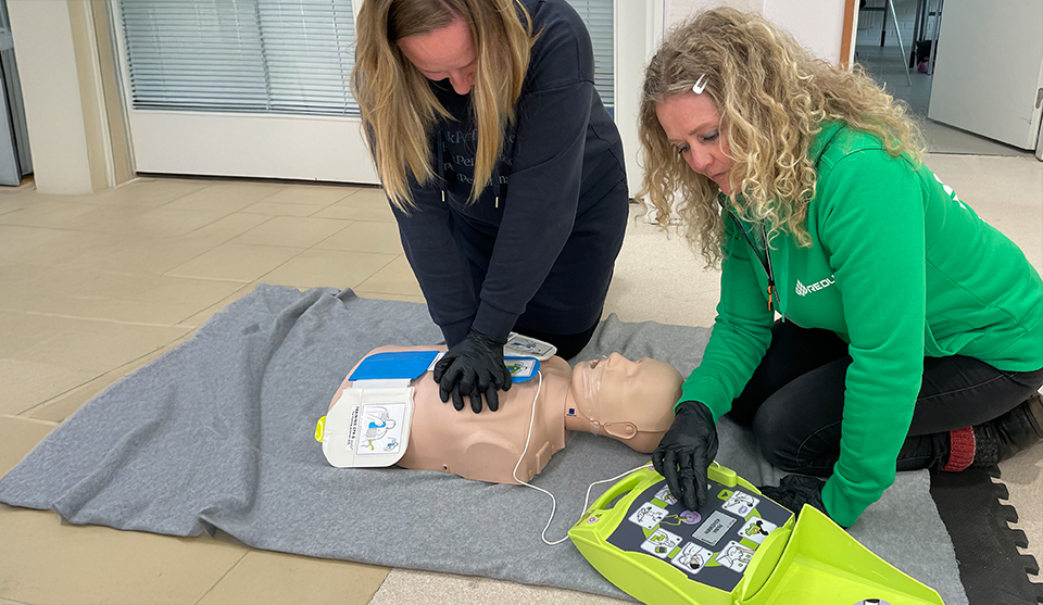 Pirjo Nissilä ja Eija Pahtakari elvyttävät nukkea ensiapuharjoituksessa. Kuvassa myös defibrillaattori.